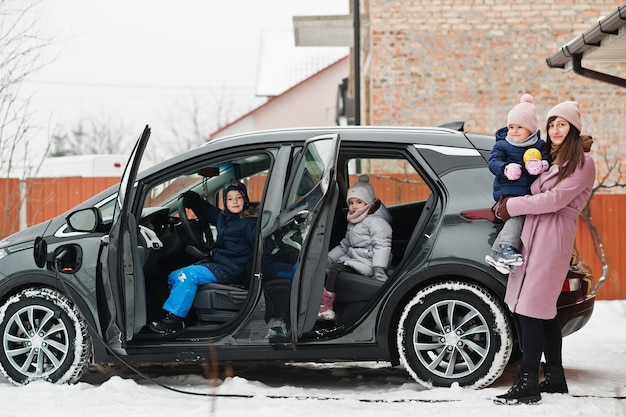 Jonge moeder met kinderen die elektrische auto opladen in de tuin van haar huis in de winter