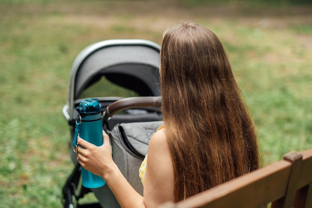 Jonge moeder met pasgeboren baby in kinderwagen drinkt water zittend op een bankje in het zomerpark jonge moeder