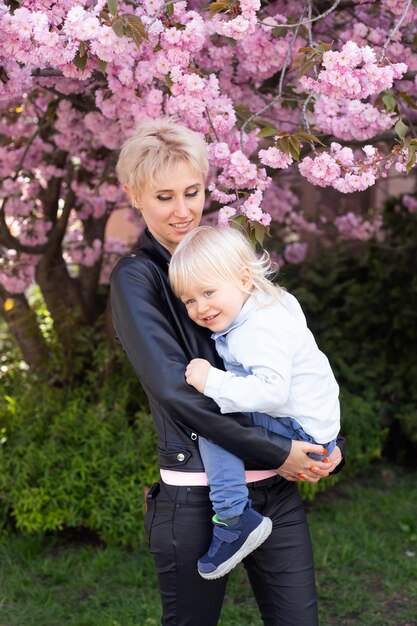 Jonge moeder moeder houdt haar zoontje van zoontje vast onder bloeiende Sakura-kersenbomen met vallende roze bloemblaadjes en prachtige bloemen