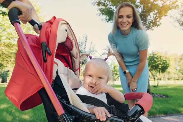 Jonge moeder wandelen in het park met lachende baby in rode zomer wandelwagen. vaders handen met kinderwagen.