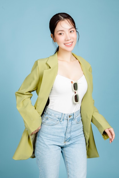 Jonge mooie aantrekkelijke Aziatische vrouw in casual groene mode jas en zonnebril geïsoleerd op pastel blauwe achtergrond Vrouwelijke consumentisme levensstijl concept