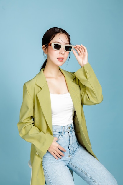 Jonge mooie aantrekkelijke Aziatische vrouw in casual groene mode jas en zonnebril zittend op een stoel geïsoleerd op pastel blauwe achtergrond Vrouwelijke consumentisme levensstijl concept