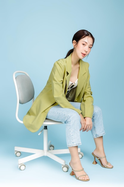 Jonge mooie aantrekkelijke Aziatische vrouw in casual groene mode jas en zonnebril zittend op fauteuil geïsoleerd op pastel blauwe achtergrond Vrouwelijke consumentisme levensstijl concept