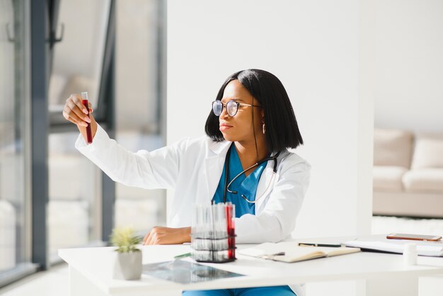 Jonge mooie African American girl arts in een witte jas met een stethoscoop. zittend aan een tafel met reagens kolven op witte achtergrond. Vrouwelijke laboratoriumassistent die bloedmonster bestudeert voor analyse