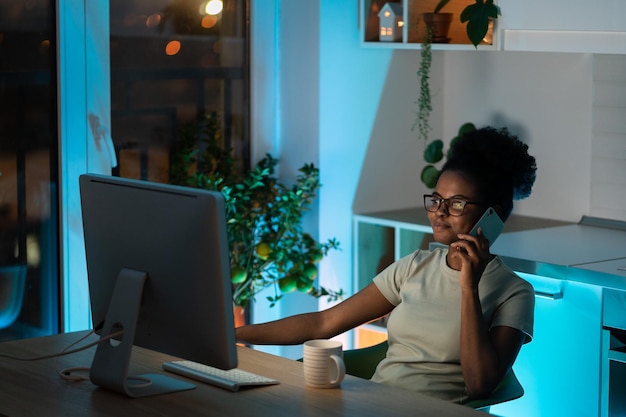Jonge mooie Afro-Amerikaanse vrouw die smartphone gebruikt terwijl ze thuis met de computer in het donker werkt