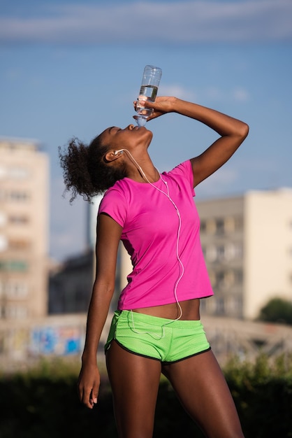 Jonge mooie afro-amerikaanse vrouw met koptelefoon drinkwater na het joggen in de stad