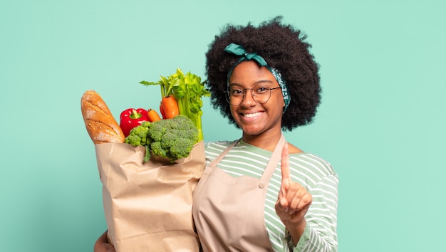 Jonge mooie afro-vrouw die zich gelukkig, opgewonden en positief voelt, een grote schreeuw geeft met de handen naast de mond, roept en een groentezak vasthoudt