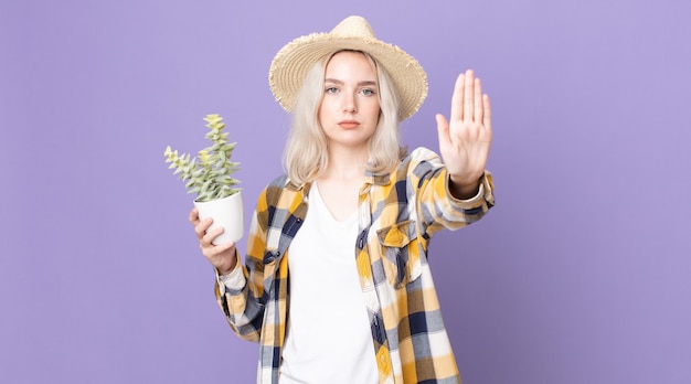 Jonge mooie albino-vrouw die er serieus uitziet en een open palm toont die een stopgebaar maakt en een kamerplantcactus vasthoudt