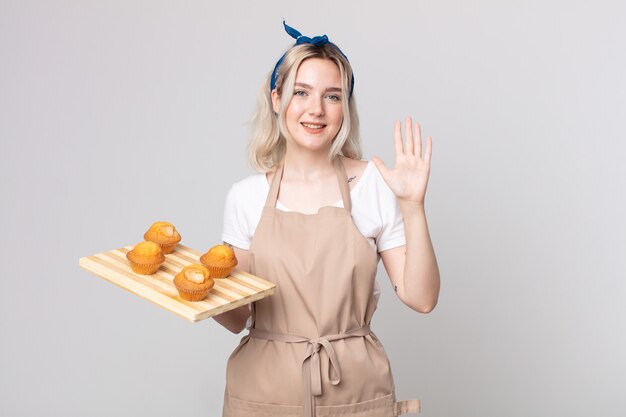 Jonge mooie albino-vrouw die lacht en er vriendelijk uitziet, met nummer vijf met een dienblad met muffins