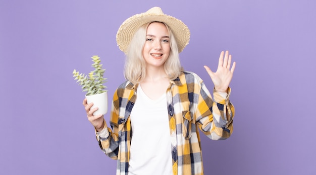 Jonge mooie albino-vrouw die vrolijk lacht, met de hand zwaait, je verwelkomt en begroet en een kamerplantcactus vasthoudt