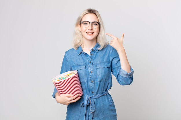 Jonge mooie albino-vrouw die zelfverzekerd glimlacht en wijst naar een brede glimlach met een emmer met popcorn