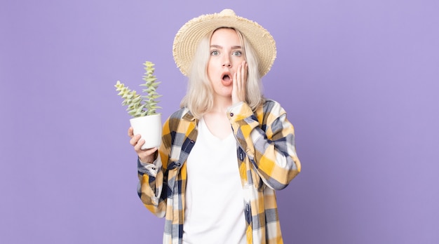 Jonge mooie albino-vrouw die zich geschokt en bang voelt en een kamerplantcactus vasthoudt