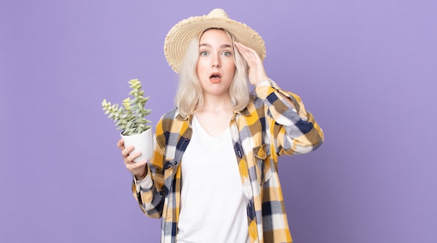 Jonge mooie albinovrouw die gelukkig, verbaasd en verrast kijkt en een kamerplantcactus vasthoudt