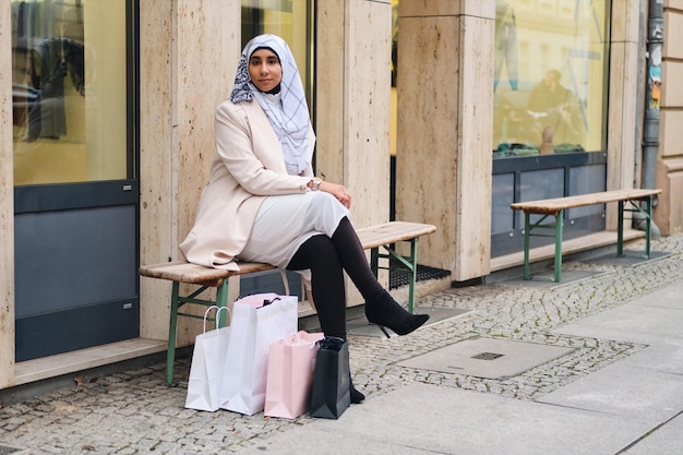 Jonge mooie Arabische vrouw in hijab rustend op de bank met boodschappentassen op straat in de stad