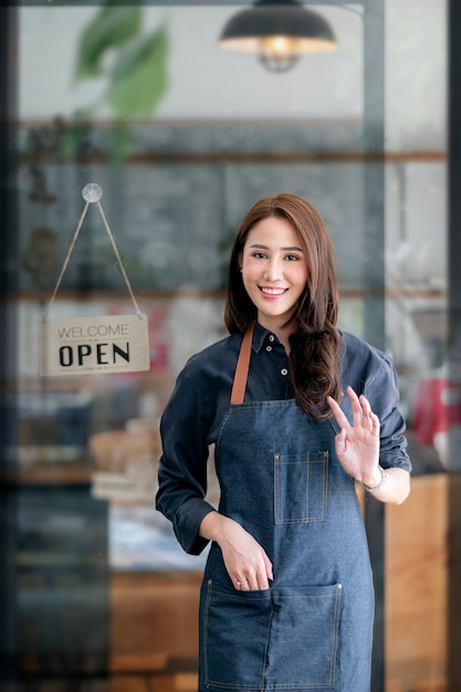 Jonge mooie aziatische barista glimlacht en kijkt naar de camera, zwaait met haar hand naar de klant terwijl ze voor de coffeeshop staat met een open bord op vensterglas.