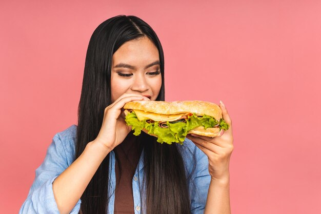 Jonge mooie Aziatische Japanse Chinese vrouw die sandwich of grote hamburger met tevredenheid eet Meisje geniet van smakelijke hamburger afhaalmaaltijden dieet concept staande geïsoleerd over roze achtergrond