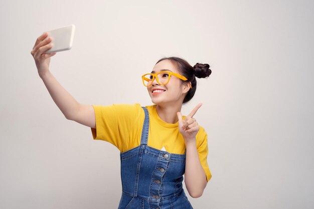 Jonge, mooie Aziatische meid die een spijkerbroek en een gele bril draagt en selfie glimlacht