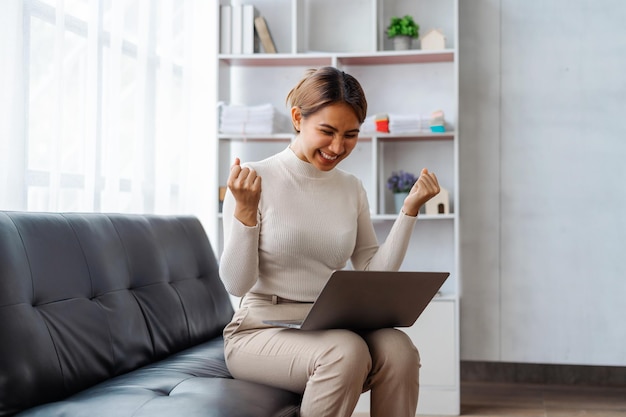 Jonge, mooie Aziatische vrouw die laptop gebruikt terwijl ze op de bank in de woonkamer zit, steekt handen op en wint overwinningsconcept