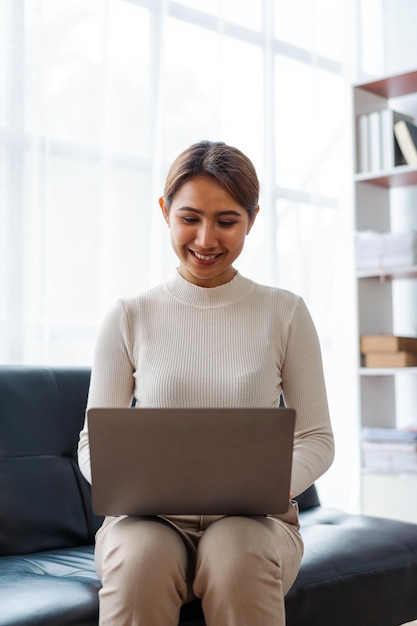 Jonge mooie Aziatische vrouw die laptop gebruikt terwijl ze op de bank in de woonkamer zit