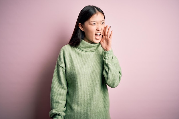Jonge, mooie aziatische vrouw, gekleed in een groene wintertrui over een roze geïsoleerde achtergrond, schreeuwend en schreeuwend naar de andere kant met de hand op de mond communicatieconcept