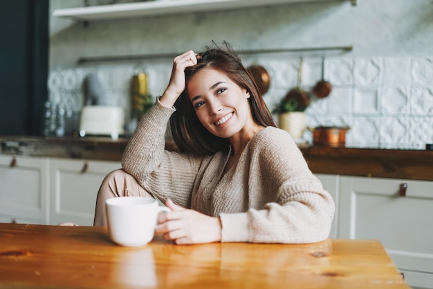 Jonge mooie aziatische vrouw in een gezellige trui zittend met een kopje thee aan tafel in de keuken thuis