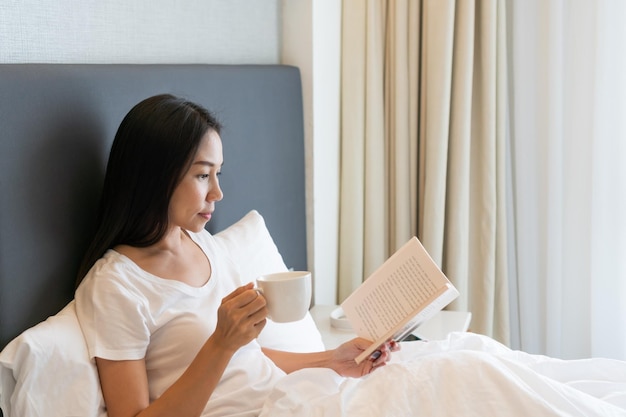 Jonge, mooie Aziatische vrouw in een witte tshirt-pyjama die koffie drinkt terwijl ze 's ochtends een boek op het bed leest Kopieer de ruimte