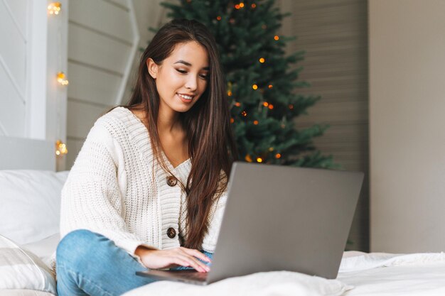 Jonge, mooie aziatische vrouw met donker lang haar in een gezellige witte gebreide trui met laptop op bed