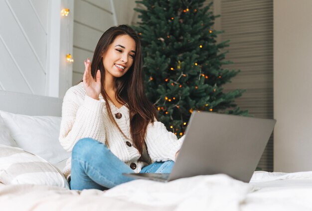 Jonge, mooie aziatische vrouw met donker lang haar in een gezellige witte gebreide trui met laptop op bed