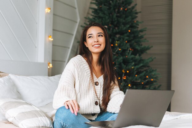 Jonge, mooie aziatische vrouw met donker lang haar in een gezellige witte trui met laptop op bed in de kamer