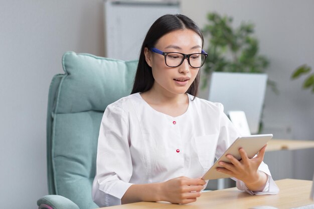 Jonge, mooie aziatische vrouwelijke arts die werkt in een moderne kliniek die tablet gebruikt voor videogesprekken en online