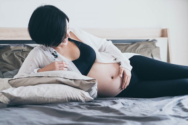 Jonge mooie Aziatische zwangere vrouw die in bed en wat betreft haar buik liggen thuis