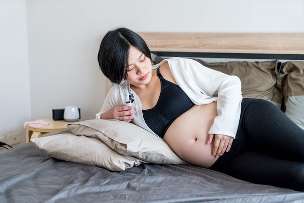 Jonge mooie Aziatische zwangere vrouw die in bed en wat betreft haar buik liggen thuis