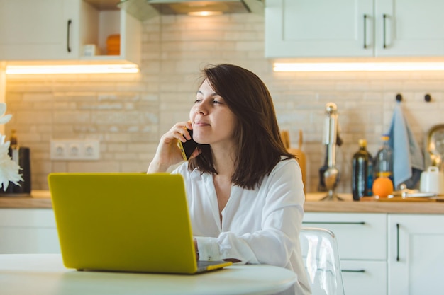 Jonge, mooie blanke vrouw die aan de telefoon praat en achter een laptop in de keuken zit