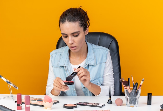 Jonge, mooie blanke vrouw die aan tafel zit met make-uptools die make-upborstel en blos vasthoudt en bekijkt