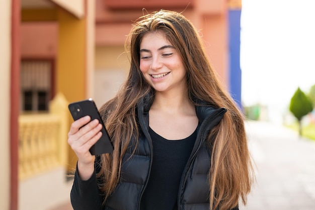 Jonge mooie blanke vrouw die buiten met een gelukkige uitdrukking een mobiele telefoon gebruikt