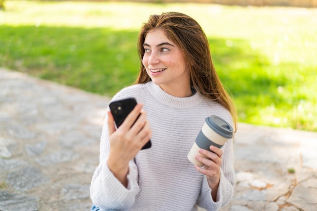 Jonge mooie blanke vrouw die mobiele telefoon gebruikt en een afhaalkoffie vasthoudt