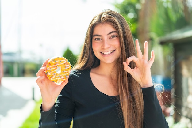 Jonge mooie blanke vrouw met een donut buiten en een ok-teken met haar vingers.