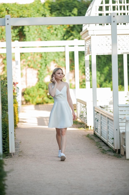 Jonge mooie blonde jaren '20 met krullend haar in een witte jurk in de zonnige zonsondergangstralen in het park Een stijlvol model loopt in de zomer door het bloeiende park van de stad