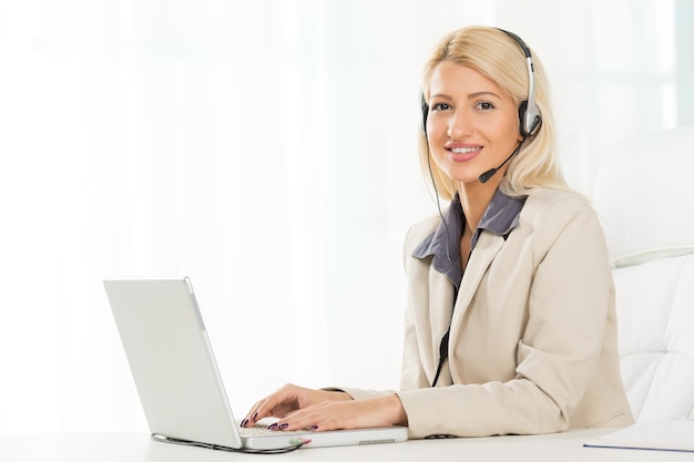 Jonge mooie blonde meid, elegant gekleed, zittend aan een bureau voor laptop met een headset op haar hoofd. Met een glimlach naar de camera kijkend.