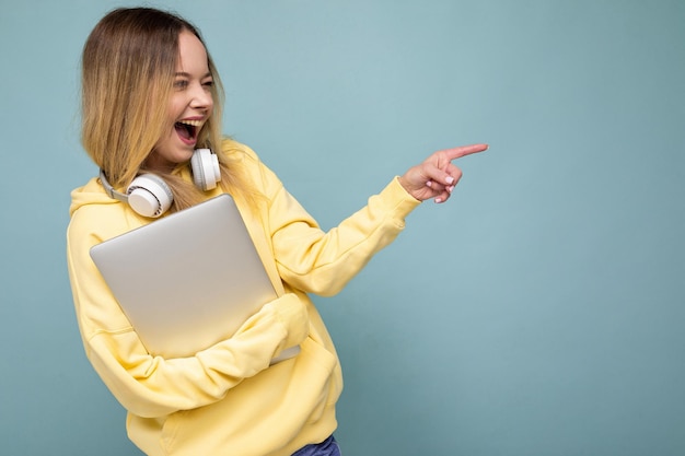 Jonge, mooie blonde student vrouwelijke persoon met een trendy gele hoodie en koptelefoon vast