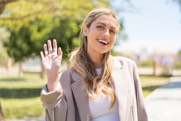 Foto jonge mooie blonde uruguayaanse vrouw buiten die met een gelukkige uitdrukking met de hand groet