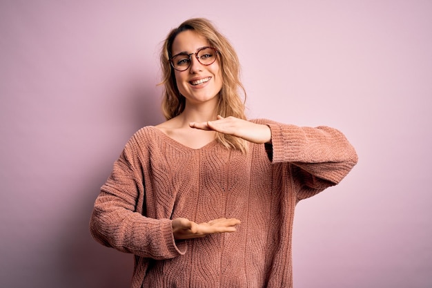 Jonge mooie blonde vrouw casual trui bril over roze achtergrond gebaren met de handen