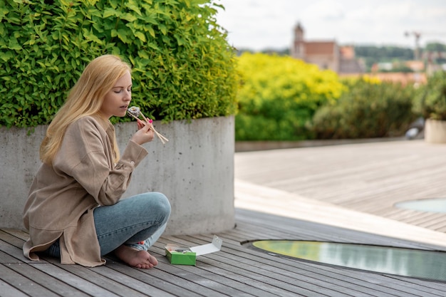 Jonge mooie blonde vrouw die buiten sushi eet op het houten terras bij modern gebouw in de stad Lekker eten om te gaan Meisje heeft lunchpauze tijd buiten doorbrengen en Aziatisch eten eten Stadsleven