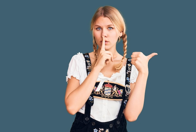 Jonge mooie blonde vrouw die oktoberfest jurk draagt en vraagt om stil te zijn met de vinger op de lippen wijzend met de hand naar de zijkant stilte en geheim concept