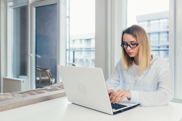 Jonge, mooie blonde vrouw die op een laptop op kantoor typt, moe van het kijken naar de camera