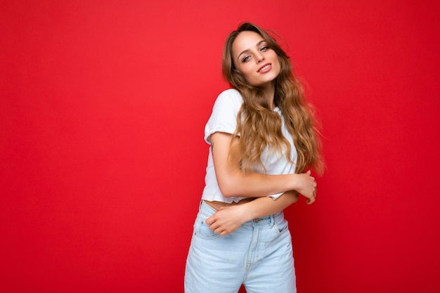 Jonge mooie blonde vrouw die wit t-shirt draagt dat camera bekijkt. Positieve vrouw toont oprechte emoties in het gezicht. Grappig model geïsoleerd op rode muur met lege ruimte