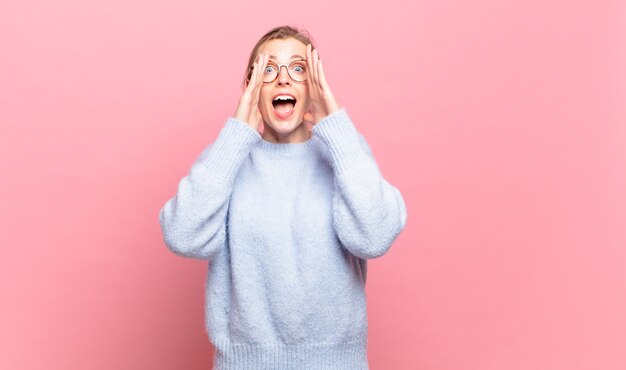 Jonge mooie blonde vrouw die zich gelukkig, opgewonden en positief voelt, een grote schreeuw geeft met de handen naast de mond, roept