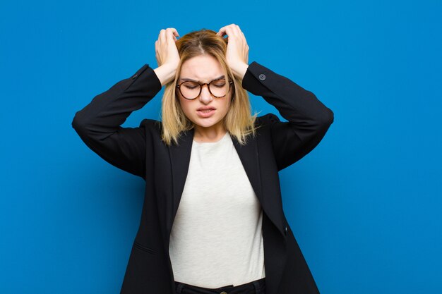 Jonge mooie blonde vrouw die zich gestrest en angstig, depressief en gefrustreerd voelt met een hoofdpijn, beide handen opheffend om tegen een vlakke muur te gaan