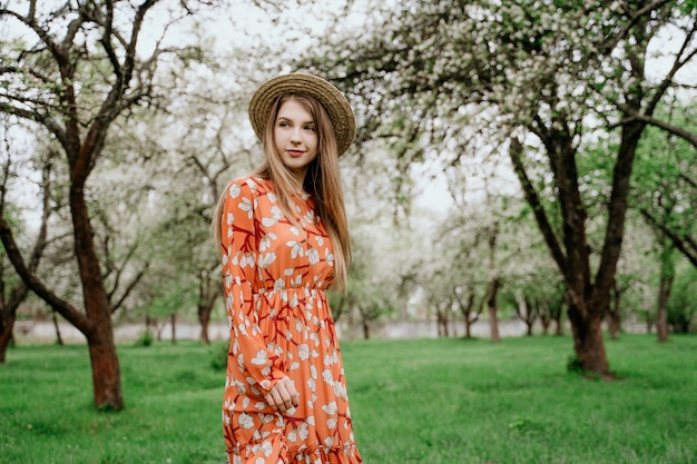 Jonge mooie blonde vrouw in bloeiende tuin. Lente bomen in bloei. Oranje jurk en strooien hoed.