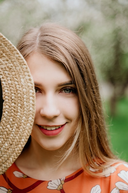 Jonge mooie blonde vrouw in bloeiende tuin. lente bomen in bloei. oranje jurk en strooien hoed.